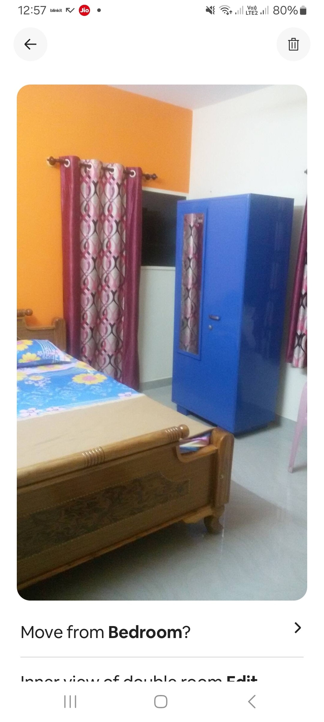 An indoor view of a double room features a spacious bed with colorful bedding. A blue cabinet stands against the wall. Bright orange and patterned curtains frame the windows, while the flooring is finished in a light tone, contributing to a calm environment.