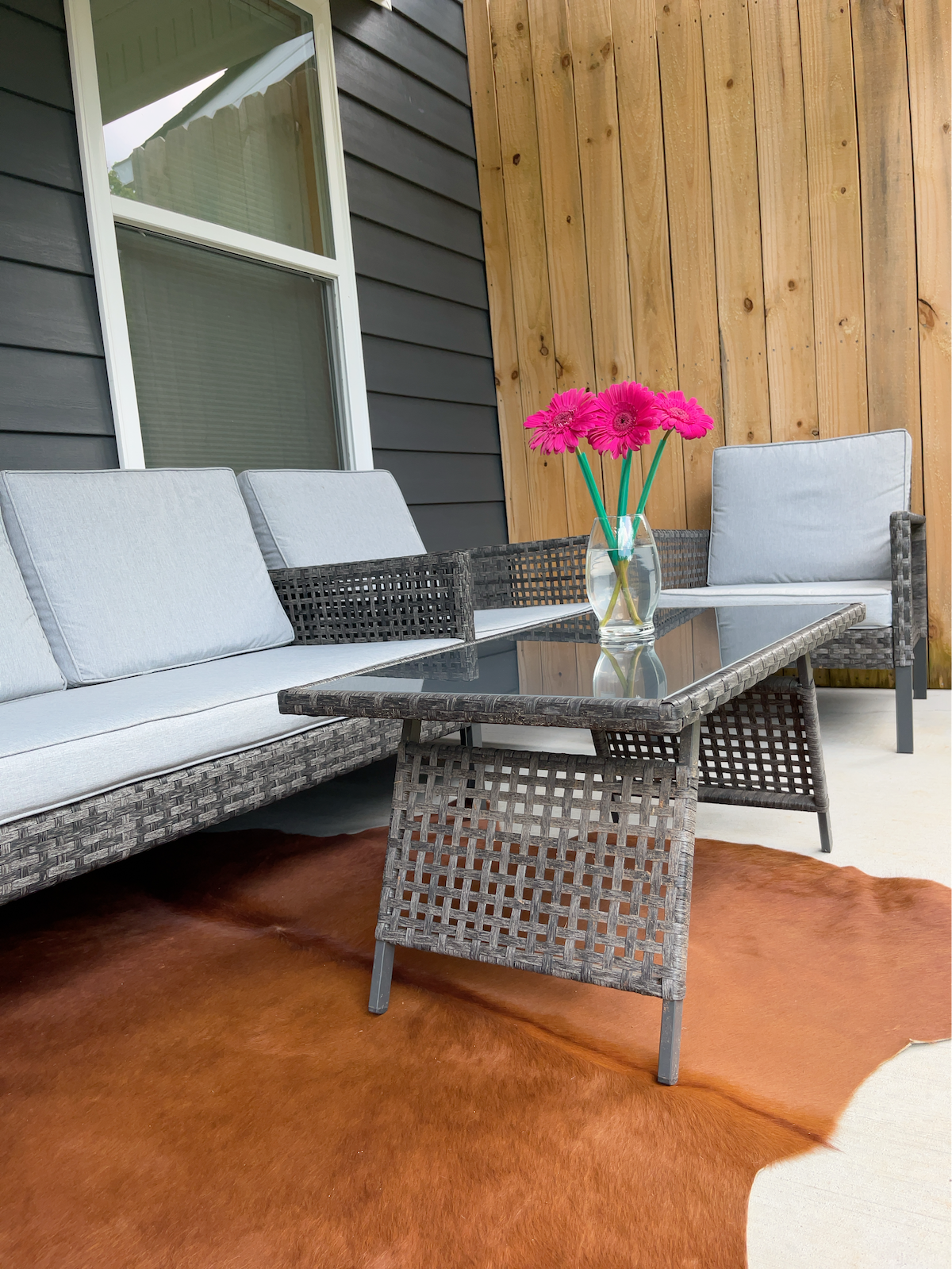 An outdoor seating area features a wicker sofa and two armchairs with soft gray cushions. A glass-topped coffee table is centered on a patterned area rug, adorned with a vase of vibrant pink flowers that adds a touch of color to the natural wood backdrop.
