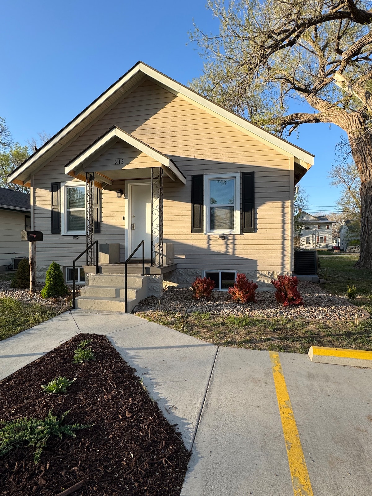 A single-story house features a welcoming front entrance with steps leading to the door. Green shrubs and small plants are lined along the path, while the exterior is adorned with beige siding and dark shutters. A clear blue sky and sunlight enhance the overall setting.