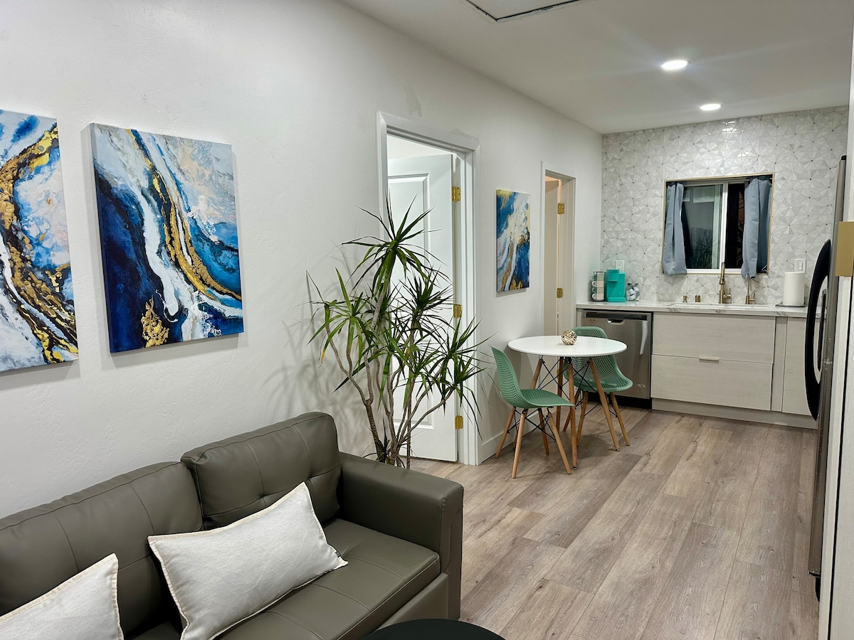 A modern living space features a green sofa with accent pillows, paired with a small round dining table surrounded by two chairs. Art pieces in blue tones adorn the walls, while a potted plant adds a touch of greenery. The kitchen countertop is visible in the background.