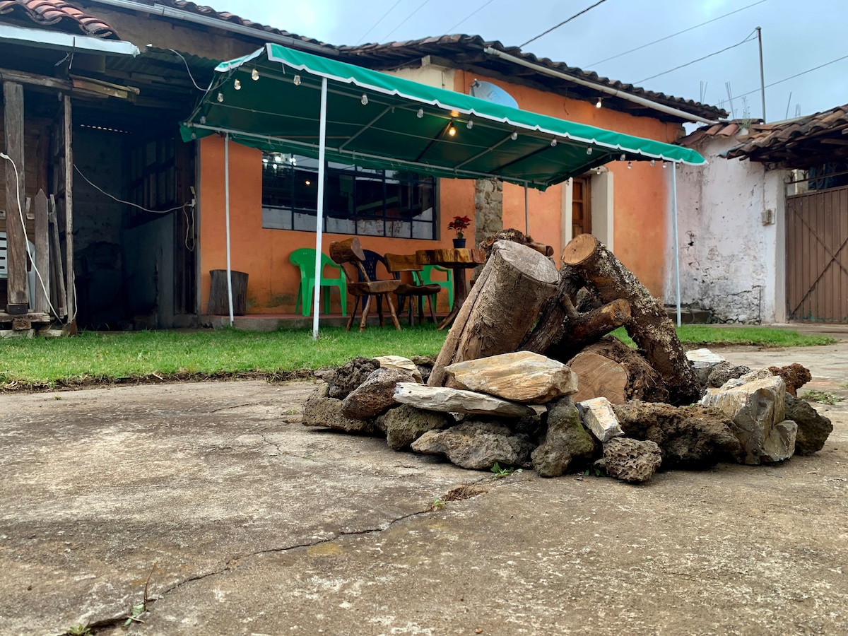 An outdoor area showcases a stone fire pit surrounded by a circular arrangement of natural materials. A covered patio is visible in the background, equipped with wooden furniture and several green plastic chairs. Soft lighting enhances the inviting nature of the space.