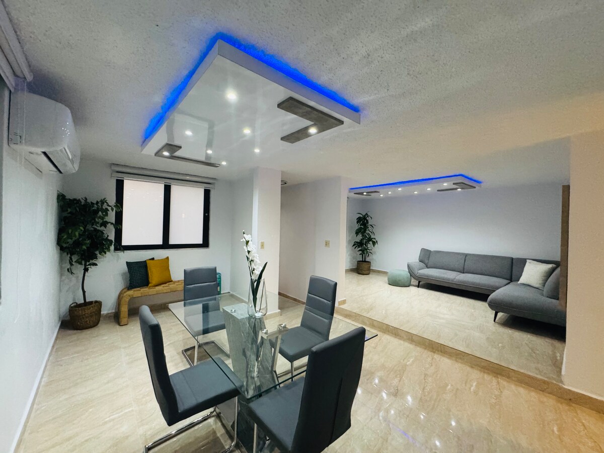 The living space features a modern interior with sleek furnishings. A glass dining table is surrounded by four black chairs, while a comfortable gray sofa is positioned in a cozy corner. Natural light enters through large windows, illuminating the marble flooring.