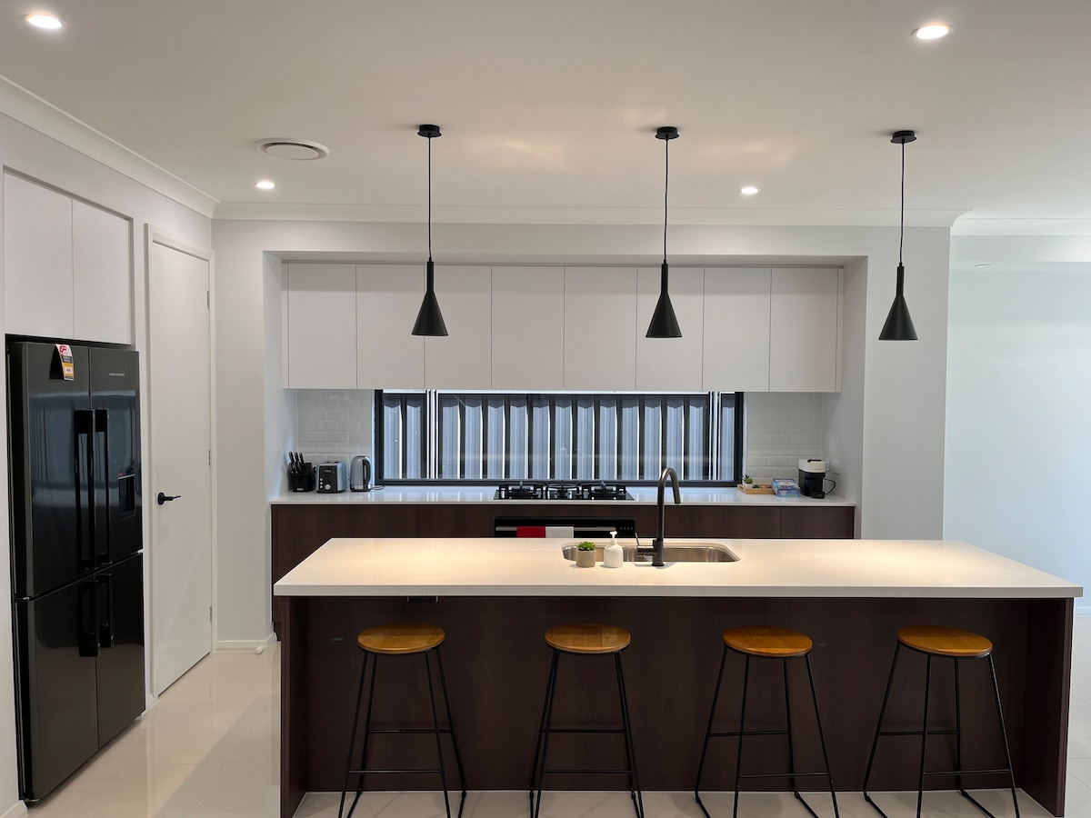 A modern kitchen features a spacious countertop with minimalist design. Three bar stools are arranged in front of a sleek kitchen island. Black pendant lights hang above, and a large refrigerator is visible on the side. The area is well-lit, emphasizing the clean lines and functionality.