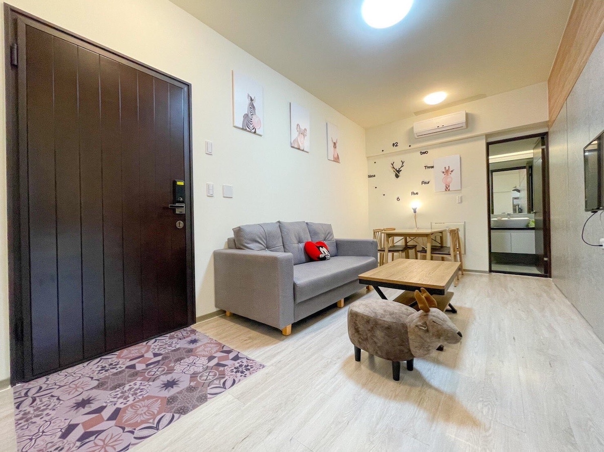 A cozy living area features a gray sofa with a decorative pillow, a wooden coffee table, and a playful stuffed animal. The light-colored walls are adorned with animal-themed artwork. A glimpse of the kitchen area is visible in the background, along with natural light streaming in.