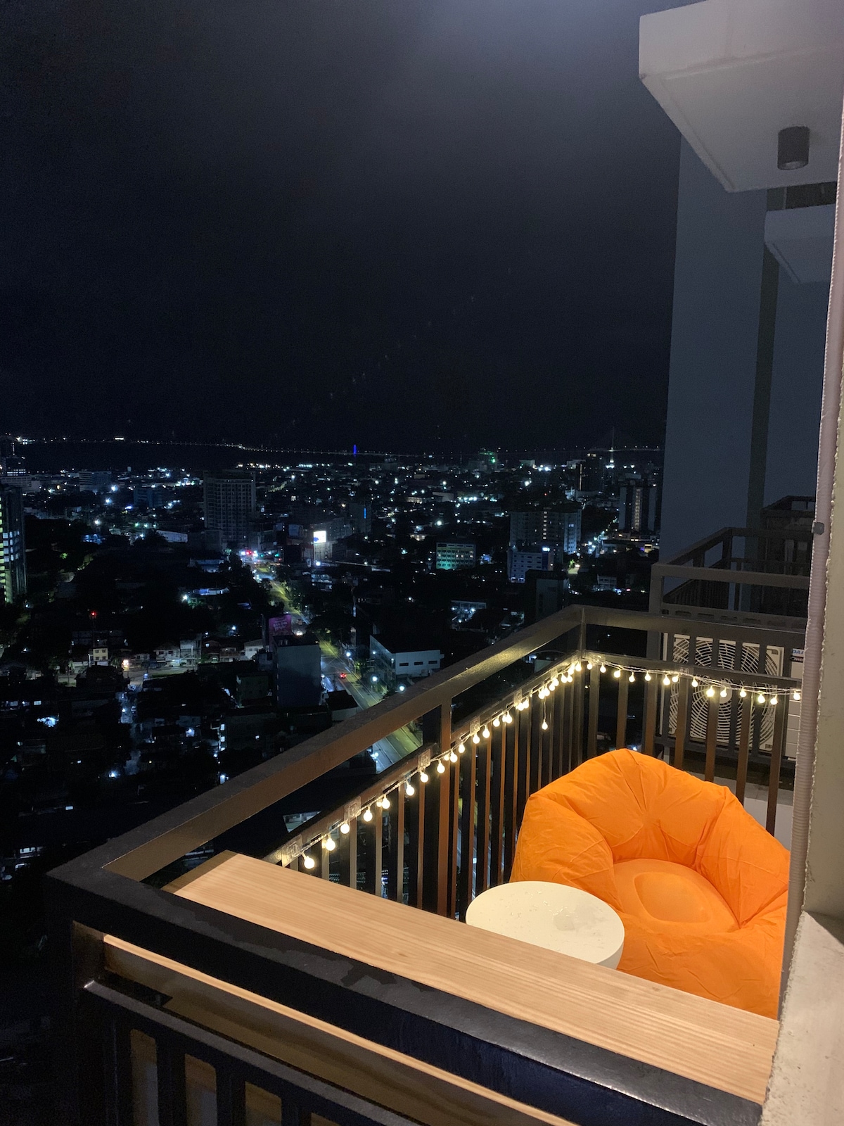 A balcony is seen at night, featuring a cozy orange bean bag and a small white table. The city skyline is illuminated with lights, showcasing a vibrant urban landscape below. String lights add a soft glow to the space, enhancing the inviting atmosphere.