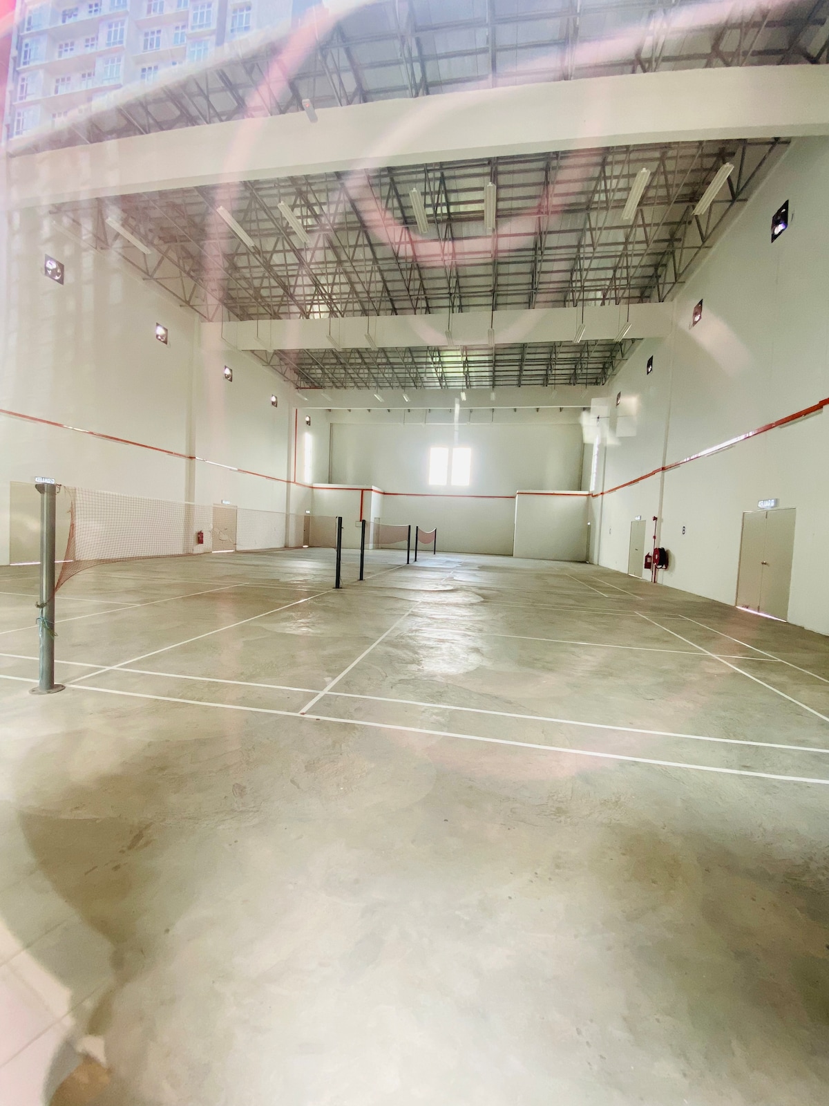 An indoor sports facility is depicted, showcasing a spacious area with a smooth concrete floor. Four badminton courts are marked with white lines. Large windows allow natural light to illuminate the space, enhancing visibility for activities.