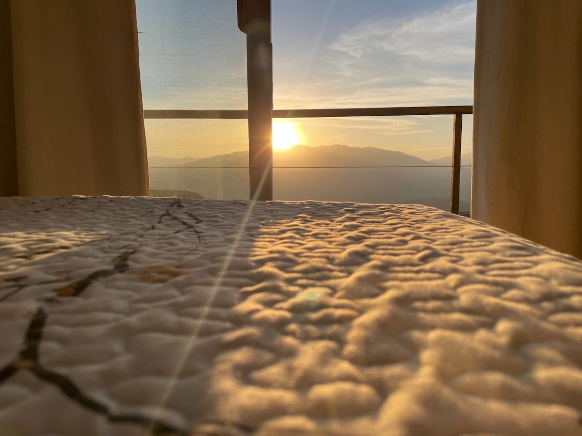 Sunrise is captured through a large window, illuminating the soft bedding with warm light. Mountains are visible in the background, offering a serene view. The sun's rays create a gentle glow across the room, suggesting a peaceful morning atmosphere.