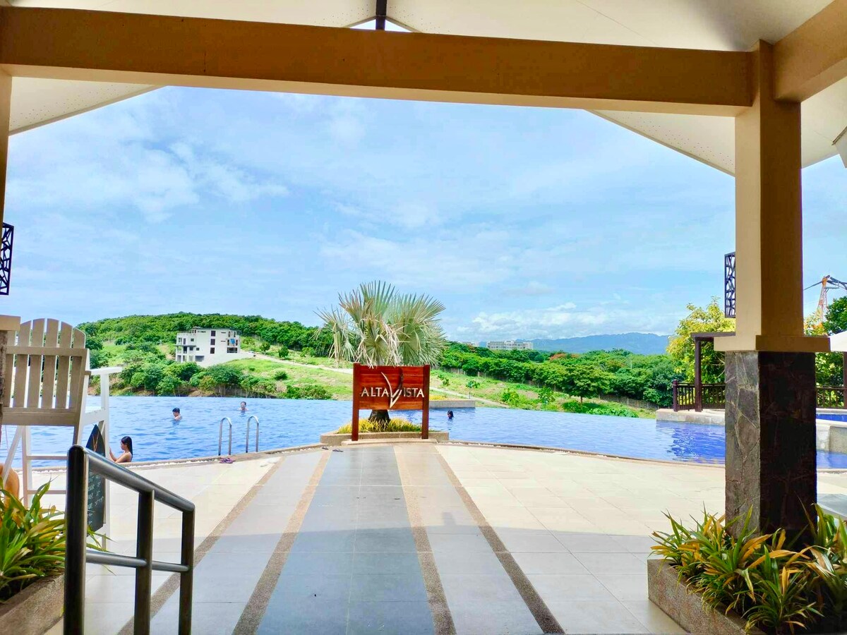 An inviting view is presented from a covered entryway, showcasing a serene infinity pool that appears to blend with the greenery beyond. Lush hills are visible in the background, framed by a bright sky, contributing to the relaxed atmosphere.