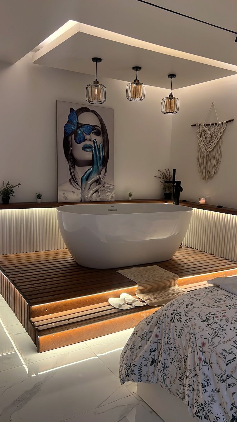 A modern bathroom features a freestanding bathtub set on a wooden platform, illuminated by soft lighting. Elegant decor, including a large art piece and woven accents, complements the serene atmosphere. A cozy bedding area is visible in the foreground, enhancing the overall design.