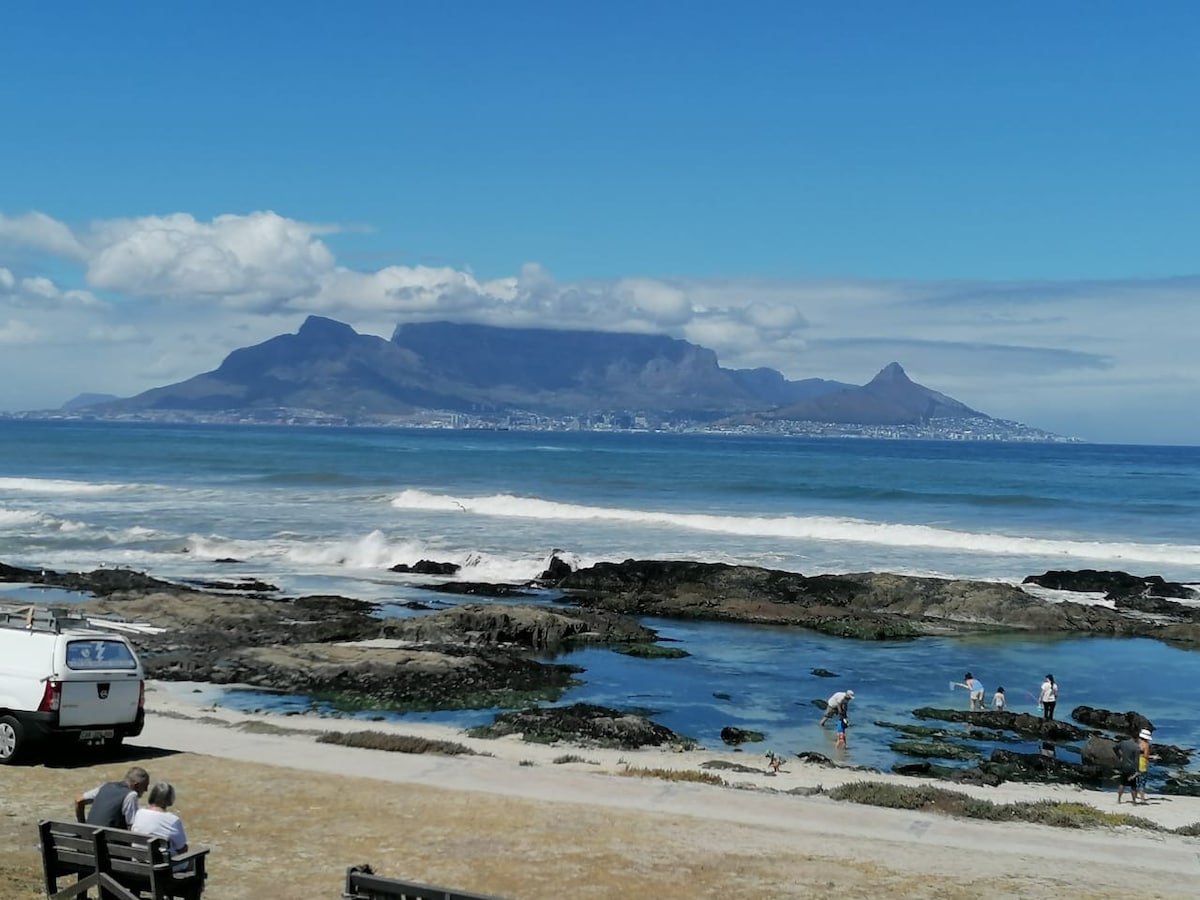 A scenic view of Table Mountain dominates the background, with waves gently crashing against rocky shores in the foreground. A couple is seated on a bench looking towards the ocean, while individuals are seen exploring tidal pools along the coastline.