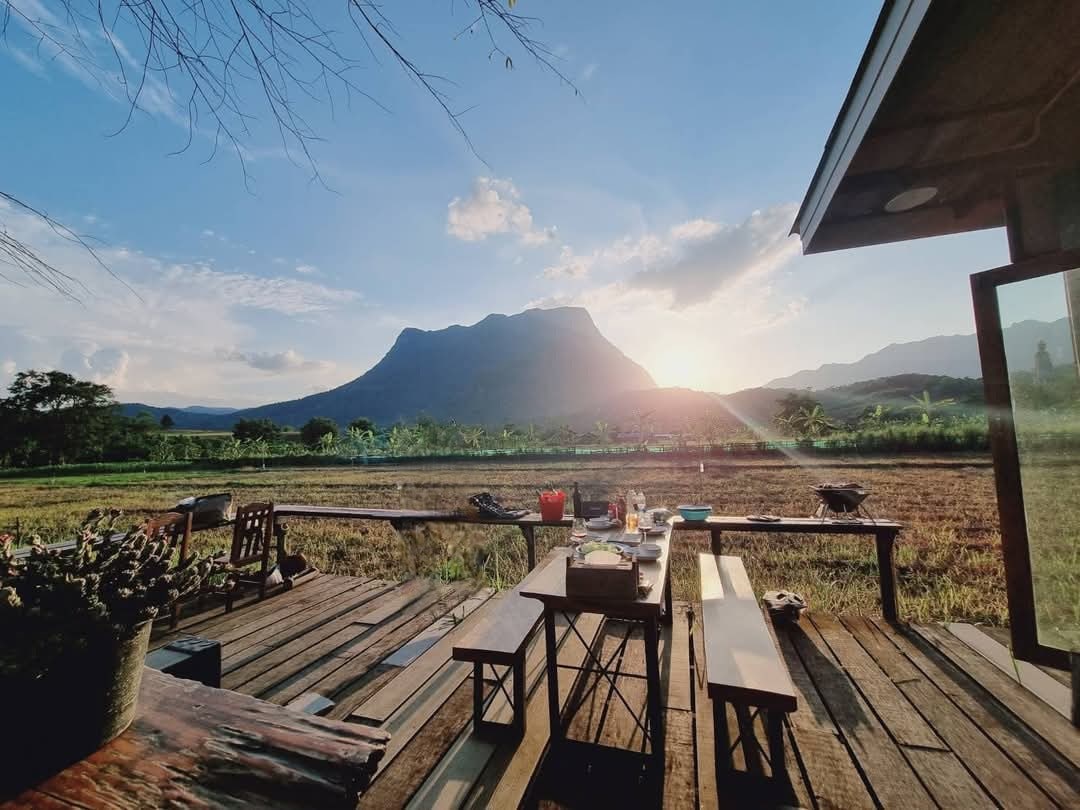 A wooden deck extends outward, offering a view of the sun setting behind Doi Luang Chiang Dao. The space features several dining tables surrounded by benches, set against a backdrop of lush fields and distant mountains.
