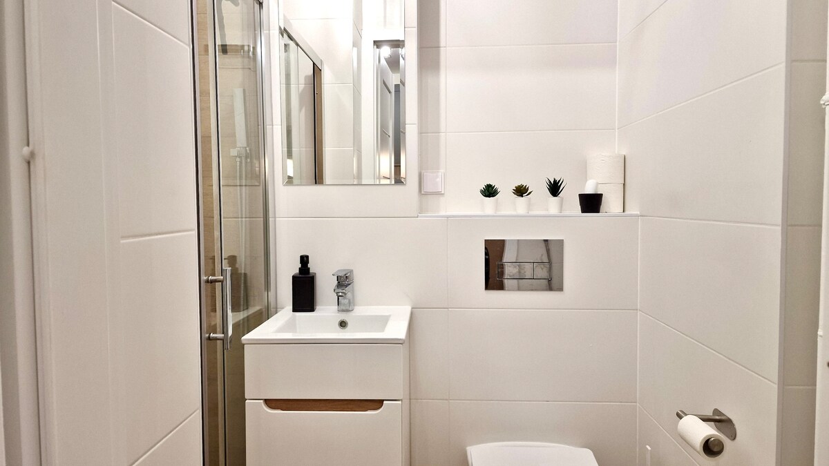 A modern bathroom is visible, featuring sleek white tiles and a glass shower. A compact vanity with an integrated sink is situated beneath a large mirror. Small potted plants add a touch of greenery to the minimalist space, which is well-lit by overhead fixtures.