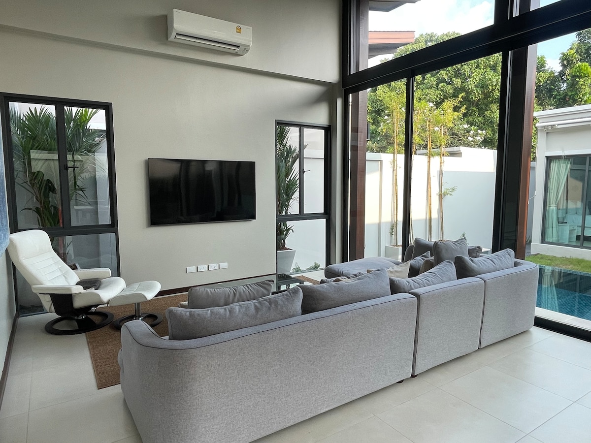 A spacious living area is showcased with large glass windows that invite natural light. A comfortable sectional sofa is arranged around a coffee table. A sleek entertainment unit holds a flat-screen television, and a white lounge chair is positioned nearby, providing a cozy spot to relax.