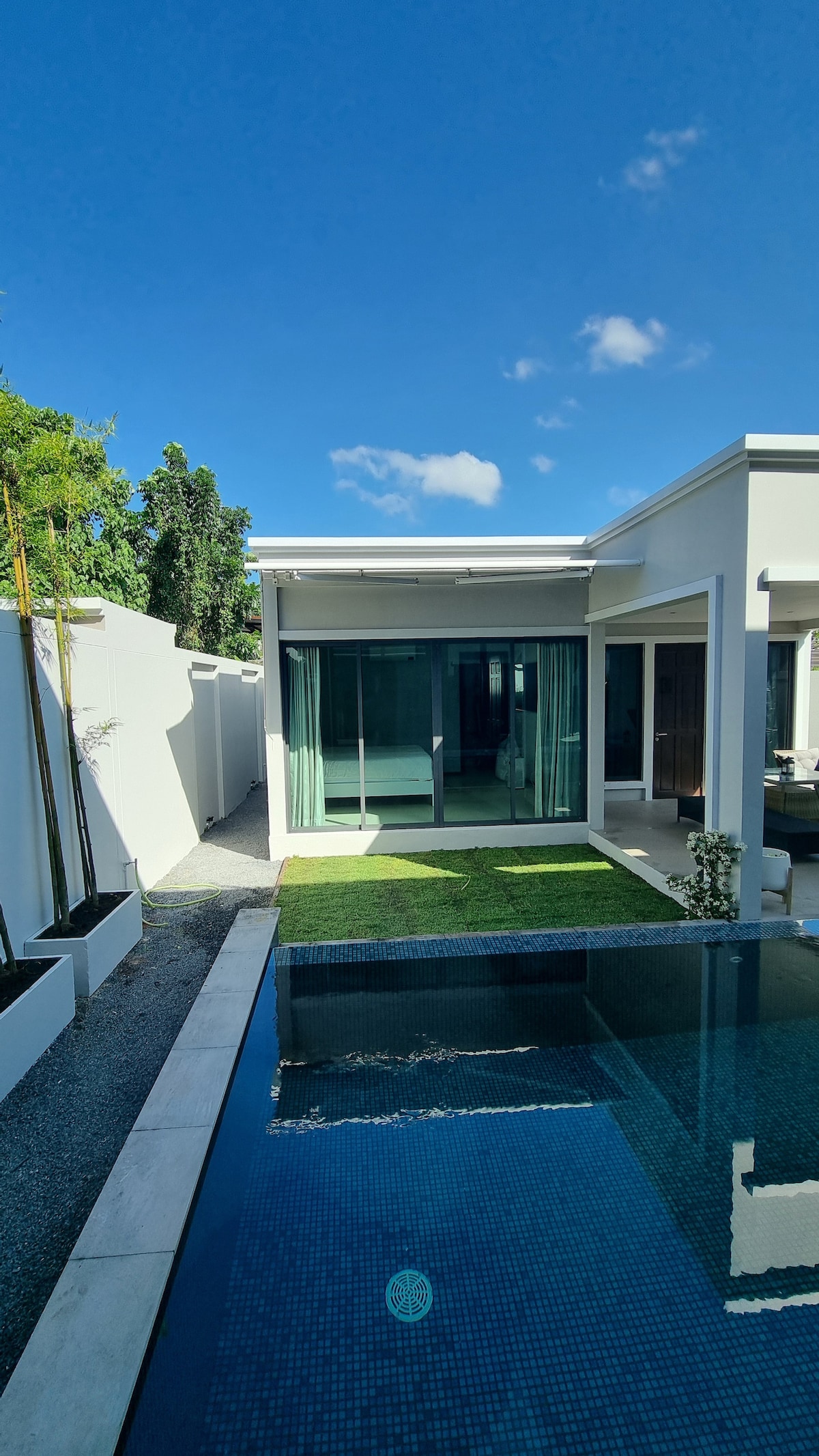 A modern villa is presented with large glass sliding doors that open to a grassy area. The space features a private pool with a tiled surface reflecting the sky. Lush greenery and minimal landscaping create a serene outdoor environment.