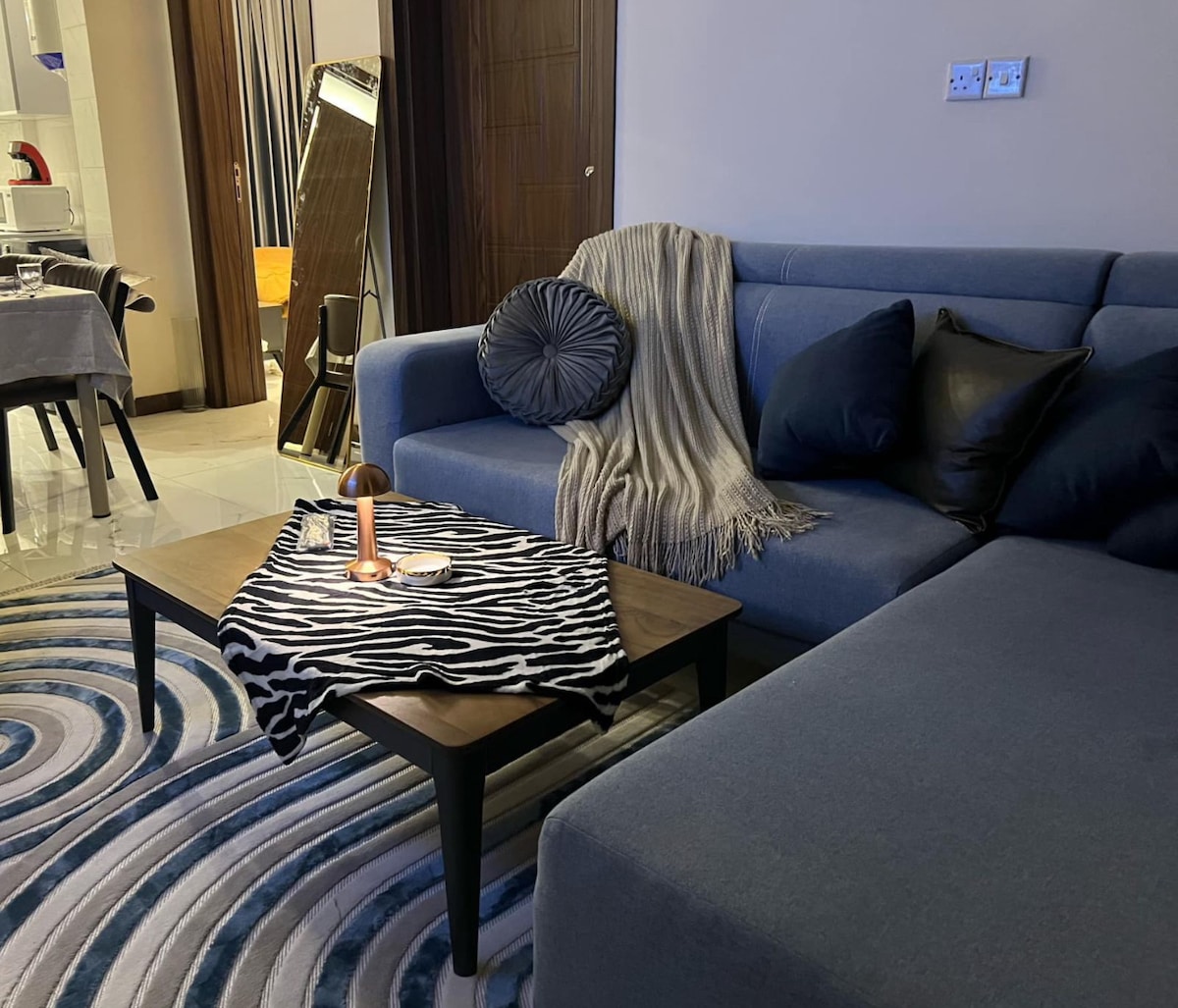A living area features a modern blue sofa adorned with plush black pillows and a textured throw. A wooden coffee table showcases a decorative zebra-print cloth, while a soft rug with circular patterns complements the space, enhancing the inviting atmosphere.
