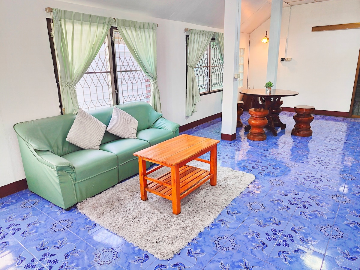 A spacious living area features a green sofa with decorative pillows, accompanied by a wooden coffee table. The floor is adorned with blue tiles, and sunlight streams through large windows, which are framed by sheer green curtains. A round dining table with wooden stools is positioned nearby.