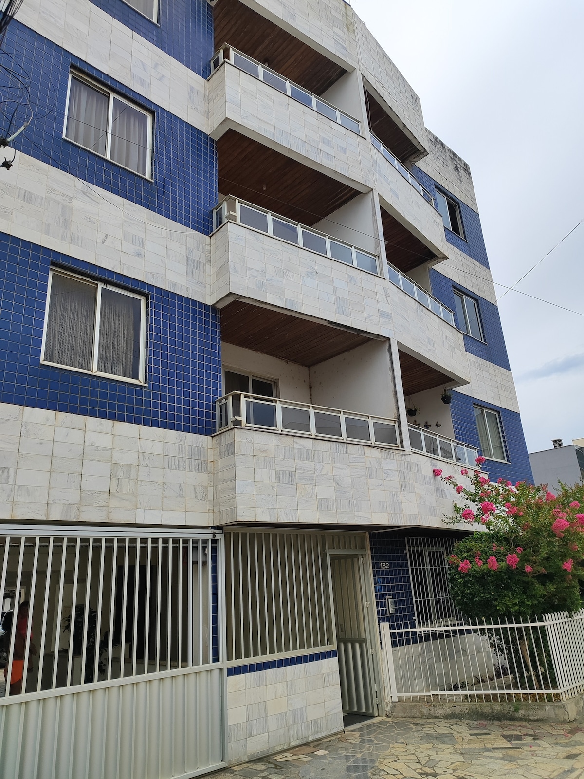 A modern three-story building features a combination of blue and white tiled exterior. Each floor has a balcony with metal railings, allowing for outdoor access. A landscaped area with flowering plants is visible at the front, and a gated entry provides security.