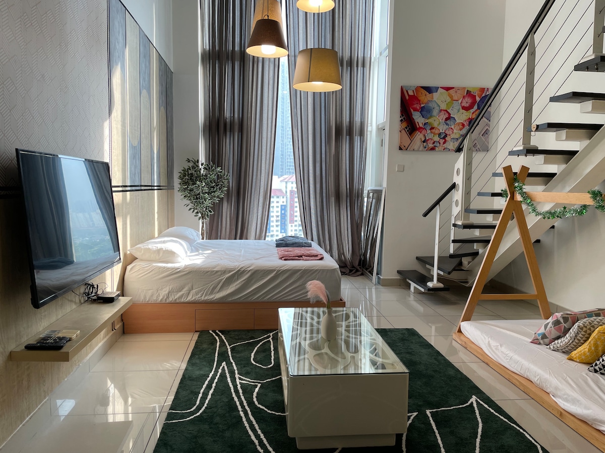 A modern living space features a spacious bed with crisp white linens and decorative pillows. Natural light filters through large windows, creating a bright atmosphere. A stylish coffee table sits on a green area rug, while a staircase and wall art add visual interest.