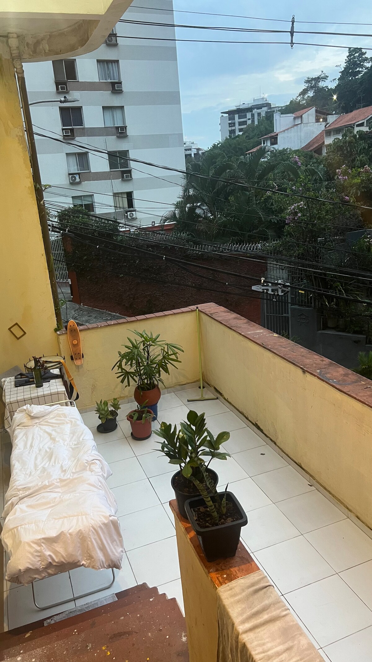 The veranda showcases a bright, tiled floor with potted plants positioned around the space. A simple table with a checkered tablecloth and a white bedspread create a relaxed atmosphere. The surrounding view includes greenery and nearby buildings, with distant mountains visible against the sky.