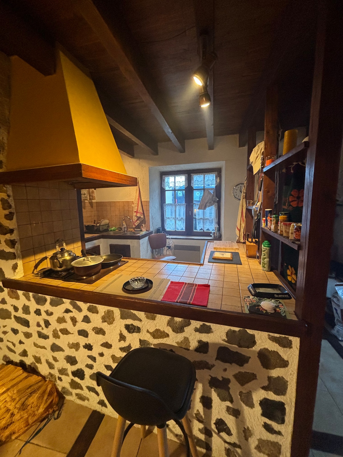 The kitchen features a warm stone wall with a light-colored tile countertop. A yellow hood rests above the stove, while wooden beams are visible overhead. A window allows natural light to fill the room, highlighting appliances and storage shelves filled with various items.
