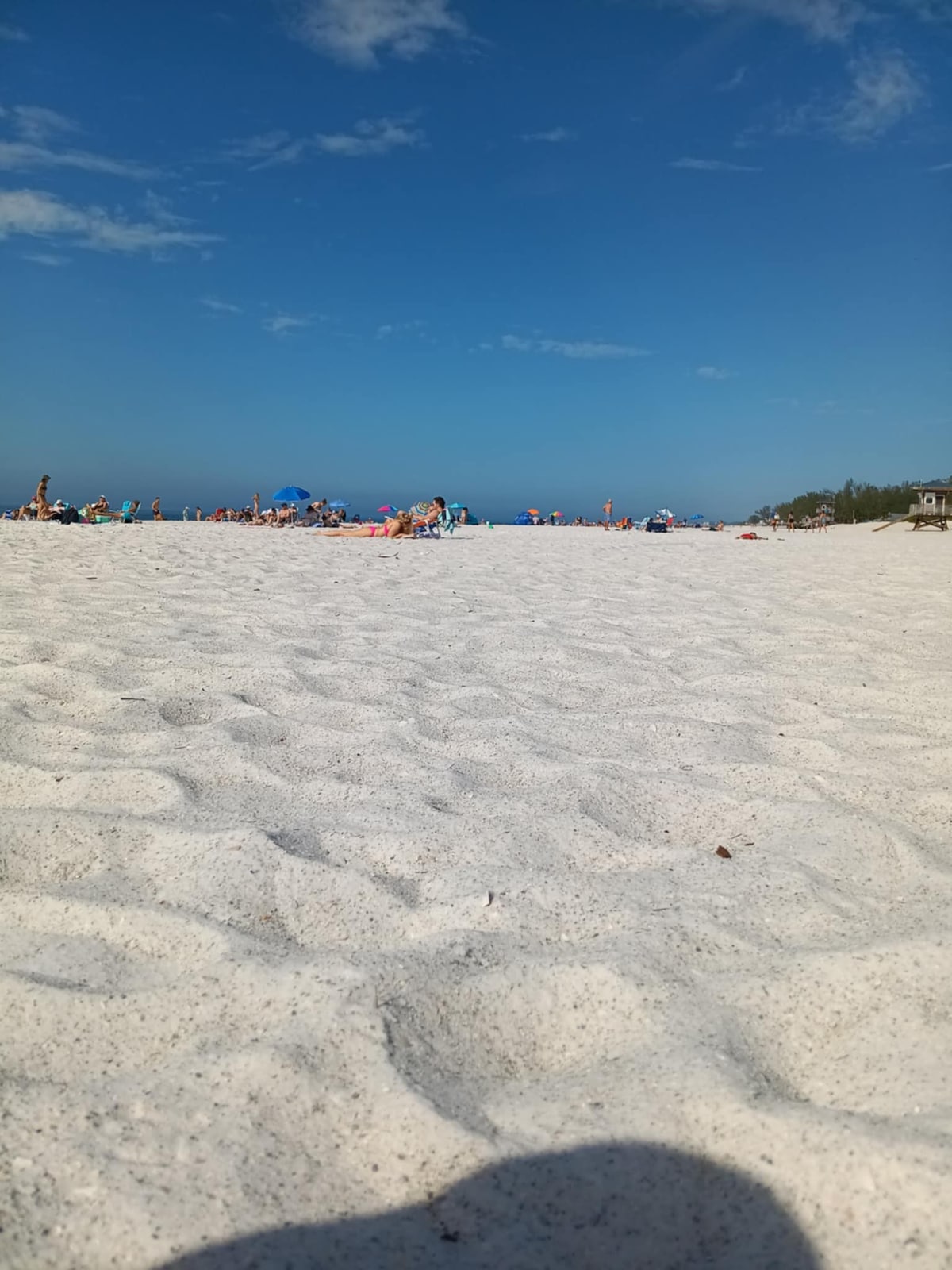 A sandy beach with soft white sand extends into the distance, dotted with colorful umbrellas and beachgoers enjoying the sunny day. The clear blue sky enhances the coastal scene, contributing to a relaxed atmosphere.
