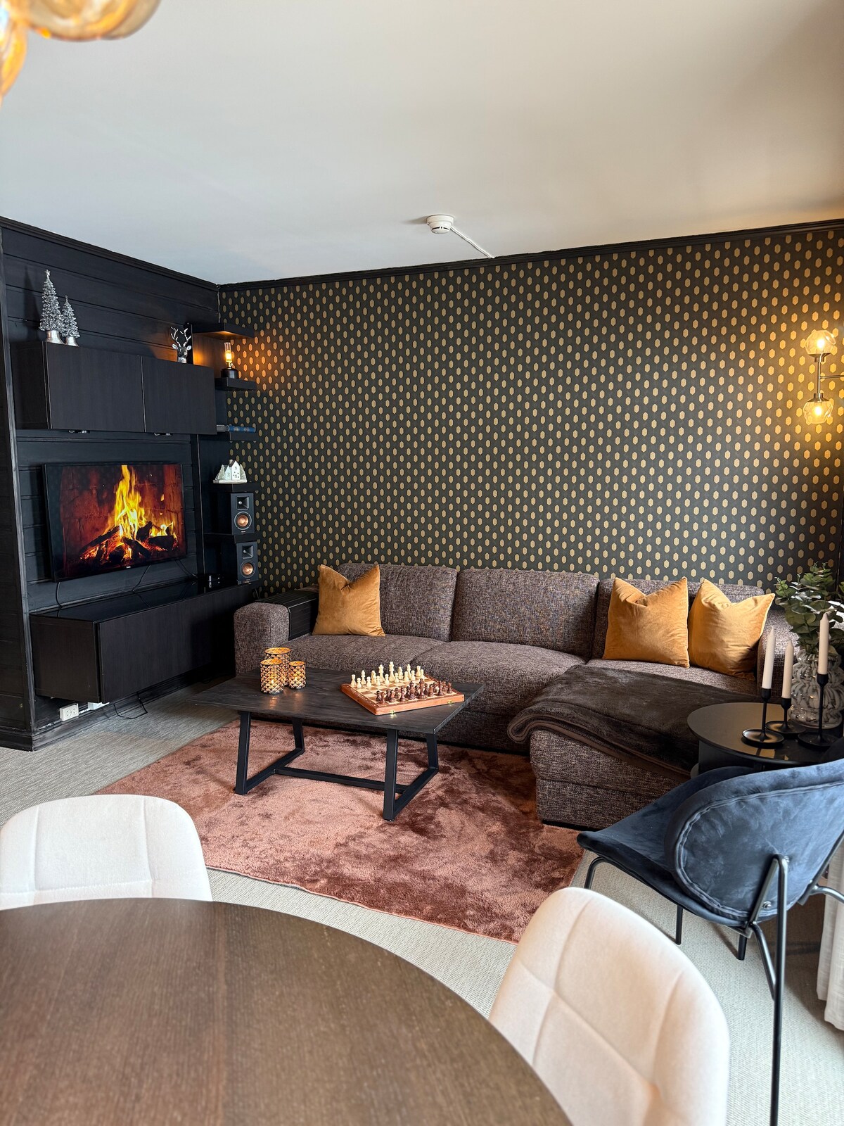 A comfortable lounge area is presented with a curved sectional sofa adorned with cushions. A decorative wall features a gold pattern, and a warm glow from wall-mounted lights enhances the space. A chess set sits on a low table, surrounded by dining chairs.