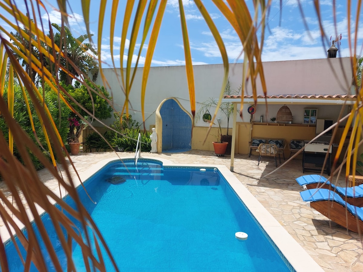 A refreshing swimming pool is framed by swaying palm leaves, with a vibrant blue surface reflecting the sky. A decorative tiled wall in the background provides a charming accent. Poolside loungers invite relaxation, alongside a shaded area that includes outdoor furniture and a barbecue space.