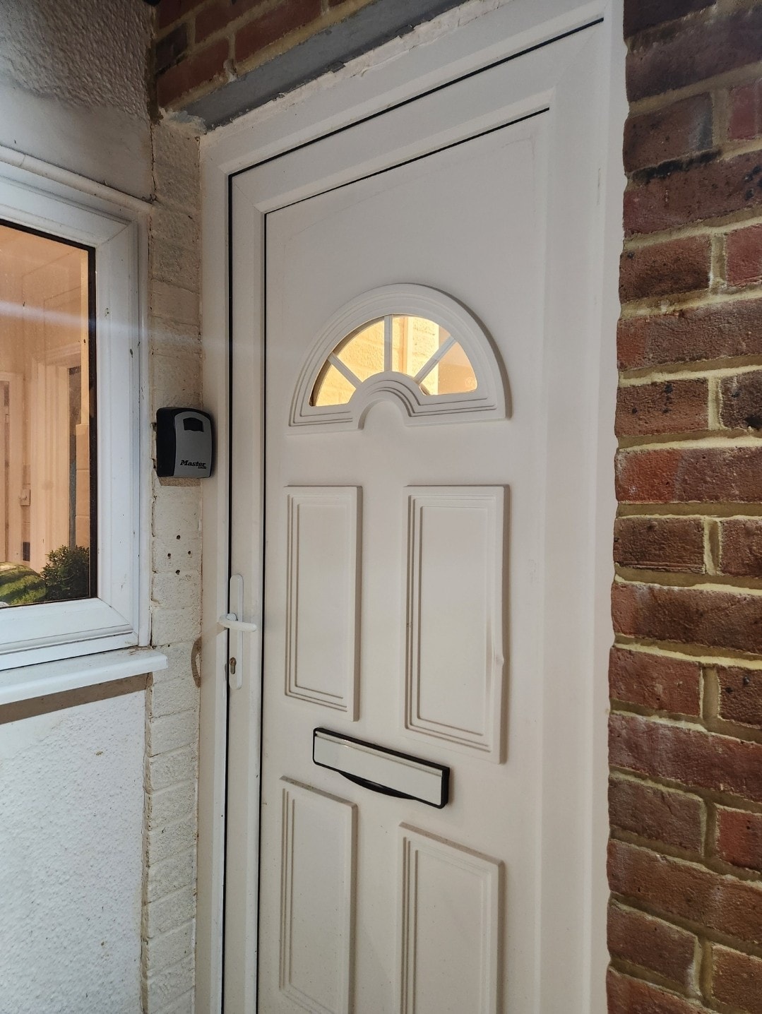 A white front door is framed by red brick walls and a nearby window. The door features an arched glass panel at the top, allowing light to filter through. An intercom is mounted beside the door, contributing to the welcoming entrance of the studio flat.