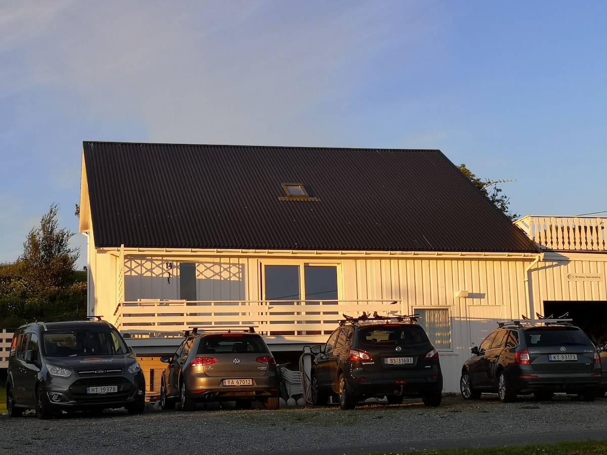 A charming two-story house features a slanted black roof and large windows, reflecting warm sunlight. Four parked vehicles are positioned in front, creating a welcoming atmosphere. A wooden balcony can be seen above a patio, enhancing the outdoor space.