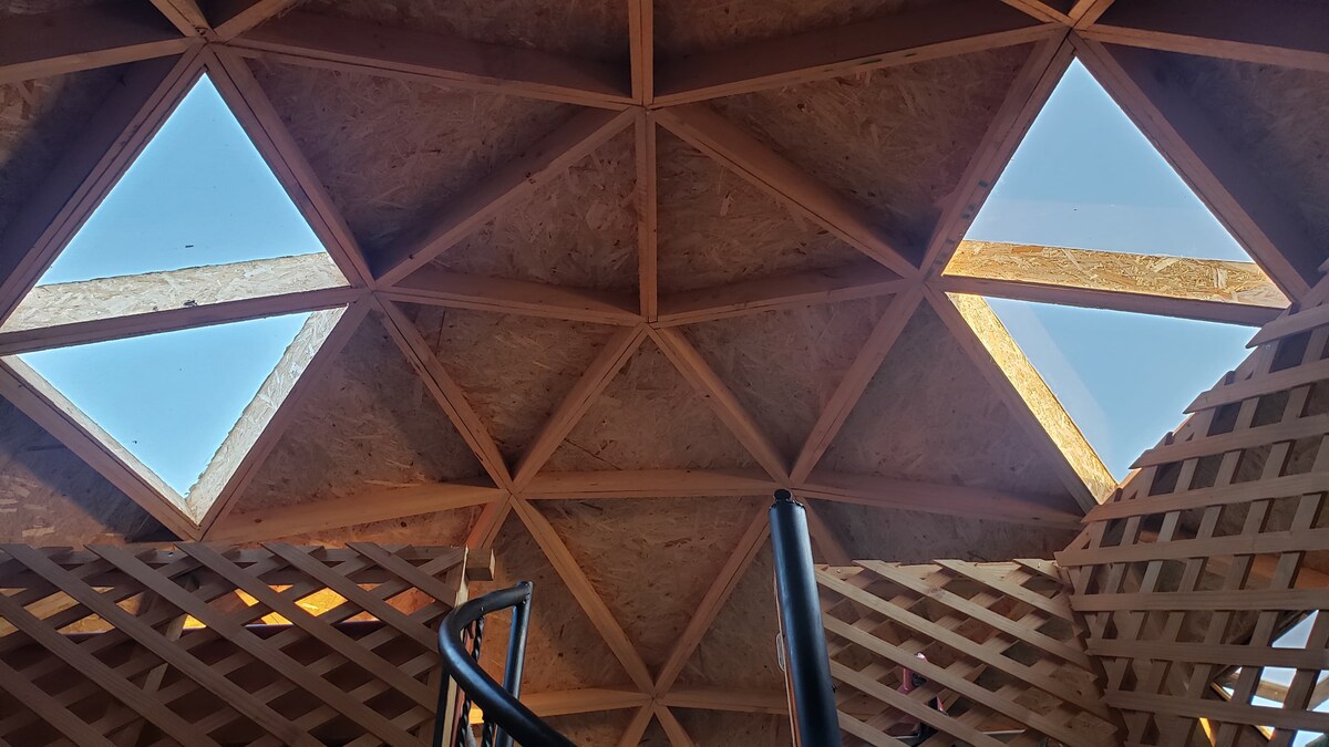 A unique ceiling design features geometric wooden beams, creating a pattern across the space. Skylights allow natural light to flood in, illuminating the interior. A spiral staircase is visible, leading to additional levels.