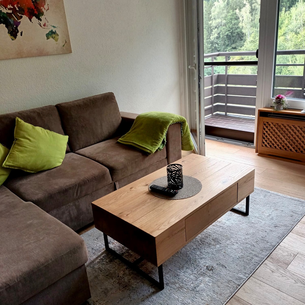 A comfortable living area is showcased with a dark brown sectional sofa accented by bright green pillows. A wooden coffee table, featuring a decorative centerpiece, is positioned on a light rug. Floor-to-ceiling glass doors lead to a balcony, revealing views of the surrounding greenery.
