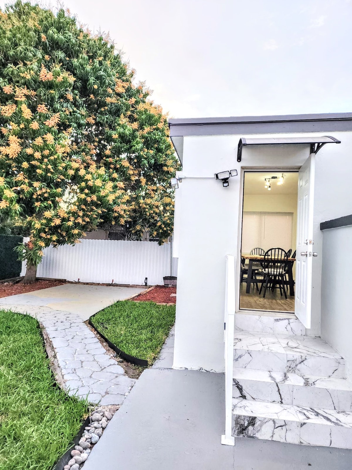 An inviting entryway is seen leading to a well-maintained path lined with stones. A mango tree provides shade in the background, alongside a grassy lawn. The exterior of the house exhibits a clean, modern design with a white door and a light fixture above.