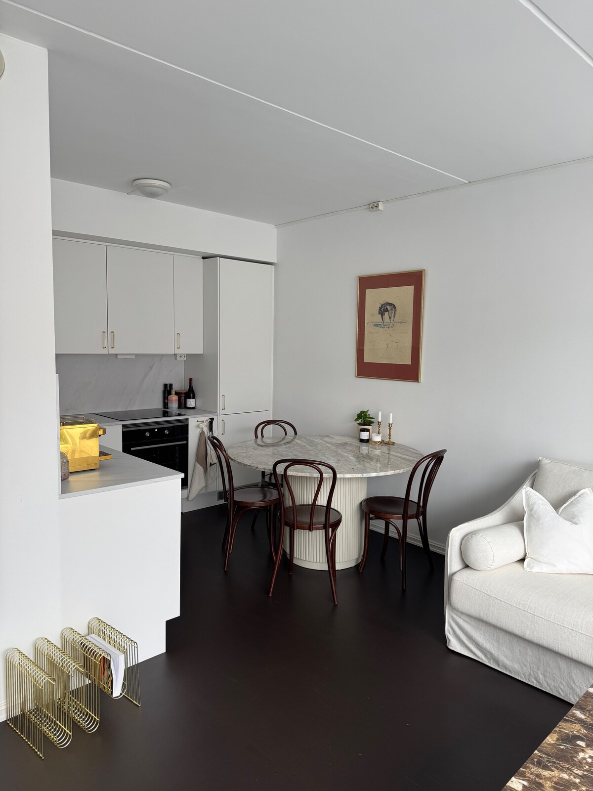 A modest dining area is visible, featuring a round table surrounded by four curved chairs. The kitchenette, equipped with modern appliances, is adjacent to the dining space. A piece of artwork is hung on the wall, adding a touch of character to the room.