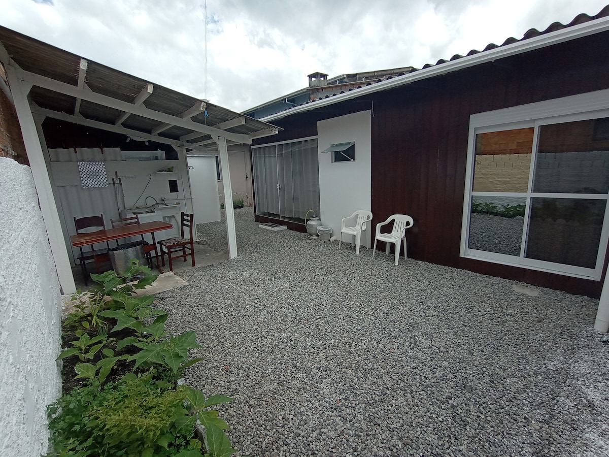An outdoor area is presented with gravel flooring, surrounded by low greenery. A shaded patio features a wooden table and chairs, while a light-colored building with large windows is visible. The space provides a serene environment for relaxation.