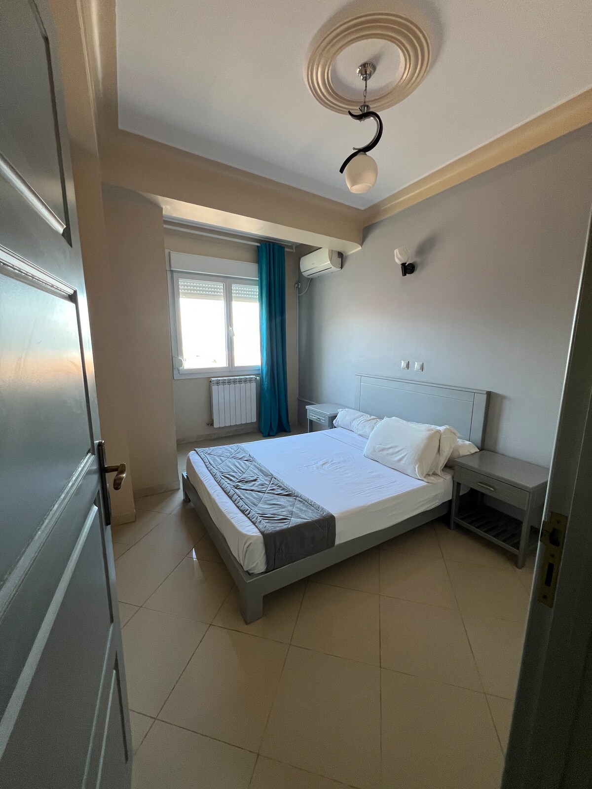 A well-lit bedroom features a spacious bed with white linens and gray accents. Natural light enters through a window adorned with light blue curtains. Two nightstands flank the bed, and a ceiling light fixture adds subtle illumination to the neutral-colored walls.