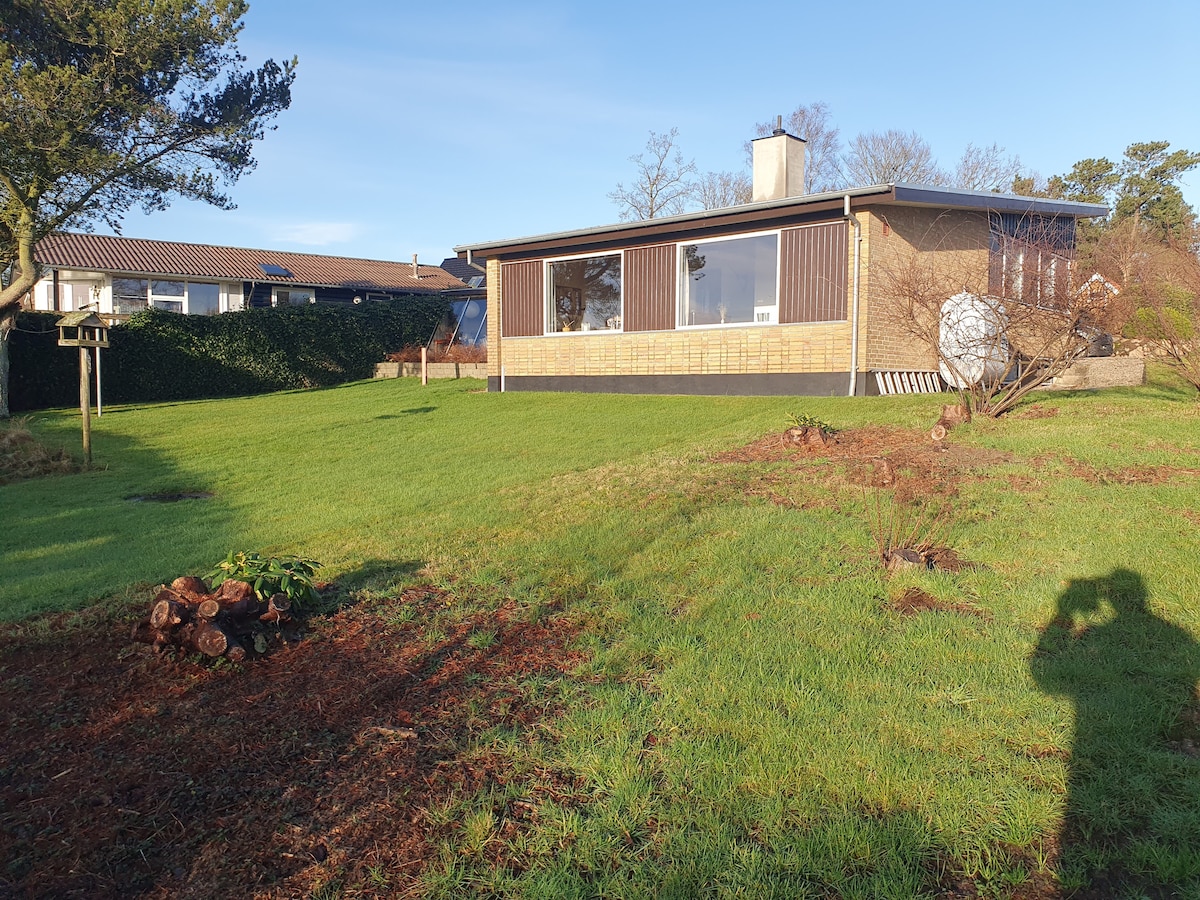 A single-story house is set on a grassy landscape, showcasing large windows that allow for ample natural light. The exterior features a combination of wooden paneling and stone elements. Nearby, a bird feeder is positioned in the yard, surrounded by greenery.