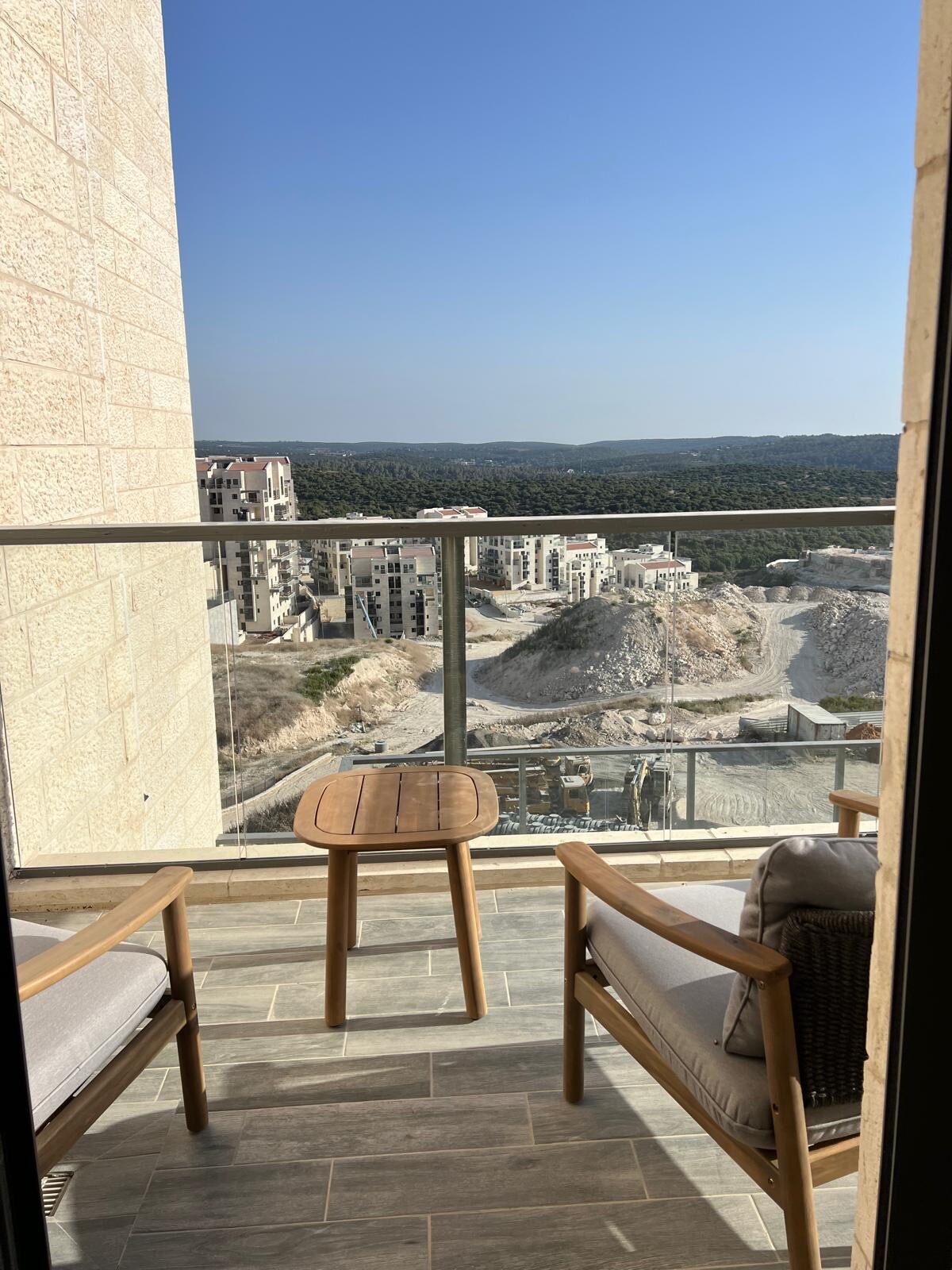 A small balcony features two wooden chairs and a round table, offering a vantage point of the surrounding landscape. The view includes hills and nearby buildings, framed by a glass railing that allows for unobstructed sightlines.