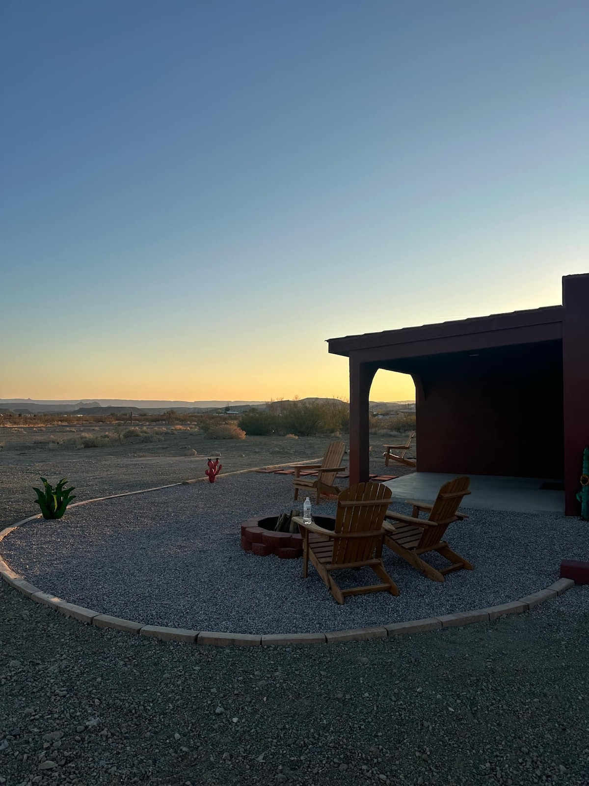 A serene outdoor seating area is presented with wooden chairs arranged around a fire pit. A gravel pathway leads to the entry of the space, which is framed by a smooth desert landscape and a clear sky transitioning into twilight.