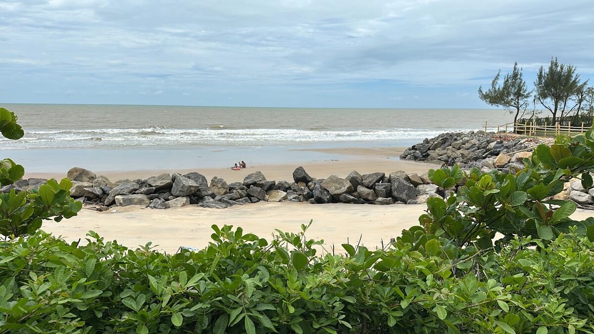 A coastal view features gentle waves lapping against the sandy shore, framed by lush green foliage. Rounded rocks create a natural barrier at the water's edge, while two figures can be seen enjoying the beach in the distance.