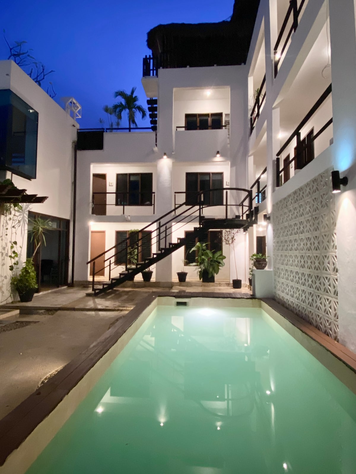 The image showcases a serene courtyard featuring a swimming pool surrounded by a light-colored structure. Soft blue lighting creates an inviting ambiance. A staircase leads to upper-level rooms, while potted plants add greenery to the space, enhancing the overall aesthetic.
