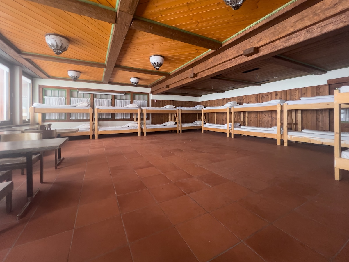 A spacious room features multiple wooden bunk beds arranged along the walls. The earthy wooden paneling complements the warm wood ceiling, while large windows allow natural light to fill the space. Simple tables are positioned to provide communal seating.