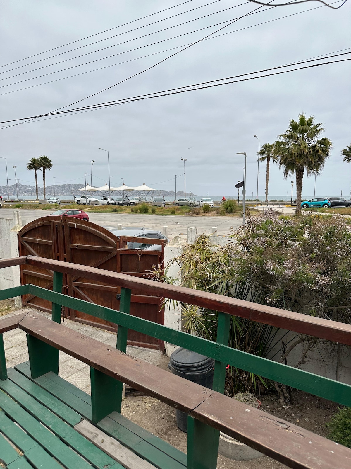 A view from a green railing offers a glimpse of the sandy beach and calm ocean beyond. Palm trees line the area, while a few distant tents can be seen under a cloudy sky. The scene is framed by a wooden gate and surrounding bushes.