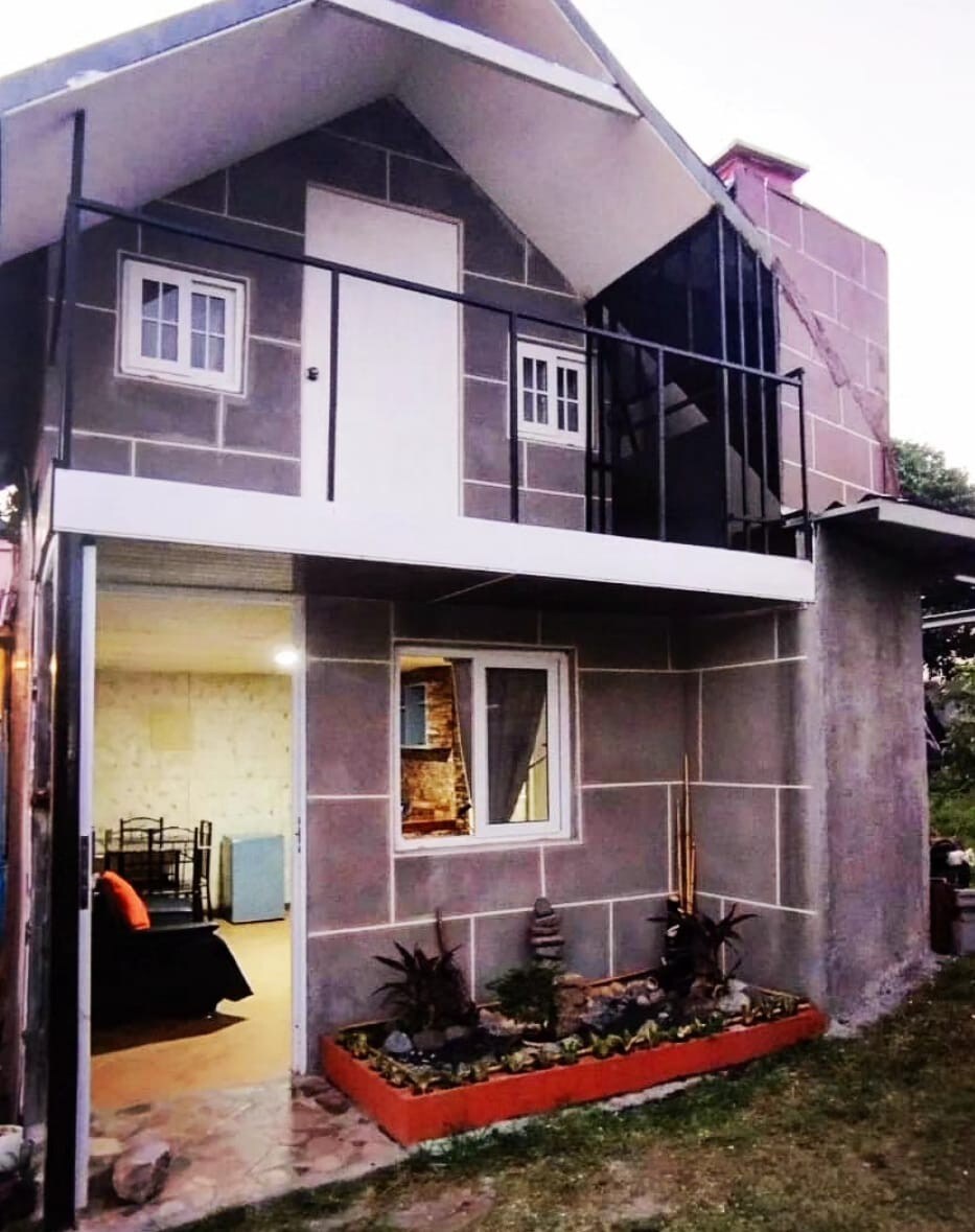 A modern two-story building is depicted, featuring a balcony with a railing on the upper level. The exterior includes a combination of wall textures, with small windows visible and a landscaped area with decorative plants in the foreground.