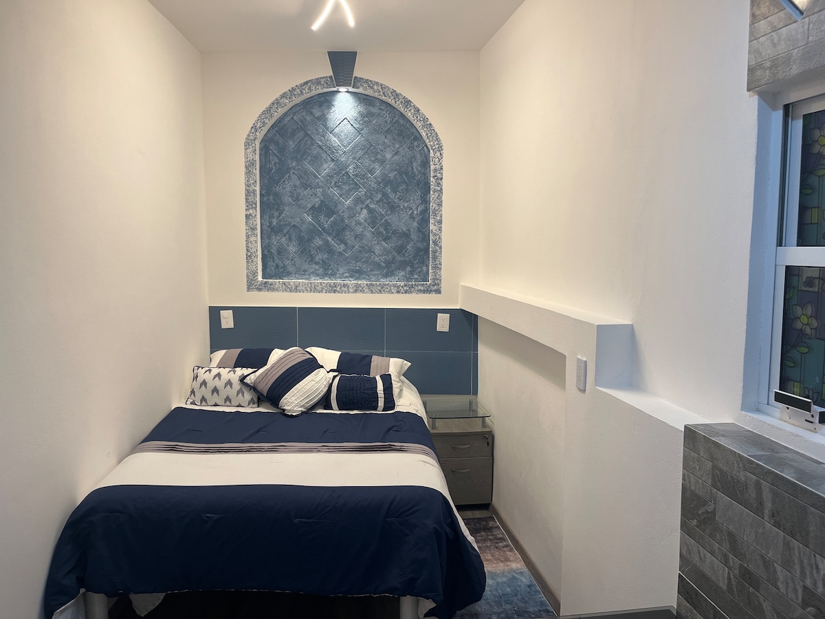 A cozy sleeping area is presented with a spacious bed adorned in navy and white bedding. A decorative arch element and a textured wall are featured above the bed. A bedside table is positioned next to the bed, and natural light enters through a nearby window.