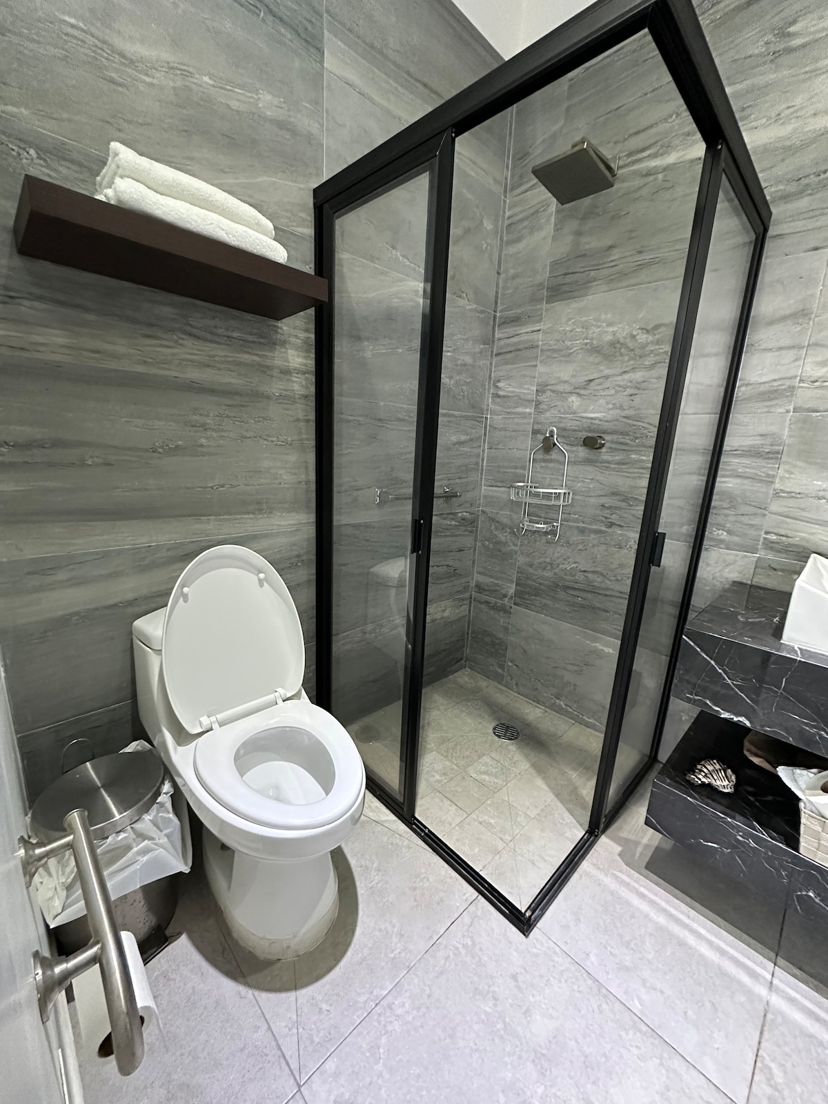A fully enclosed bathroom is shown, featuring a glass shower stall with a rainfall showerhead, a modern toilet, and a wall-mounted shelf with neatly folded towels. The space is adorned with marble and granite finishes, enhancing the sleek design.