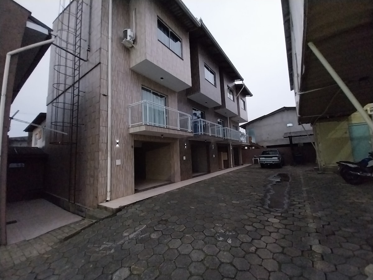 A two-story residential building is depicted, featuring a light-colored facade with balconies on each upper unit. An outdoor parking area is visible, along with a smooth stone pathway. A car is parked nearby, while shaded spaces can be seen under the structure.