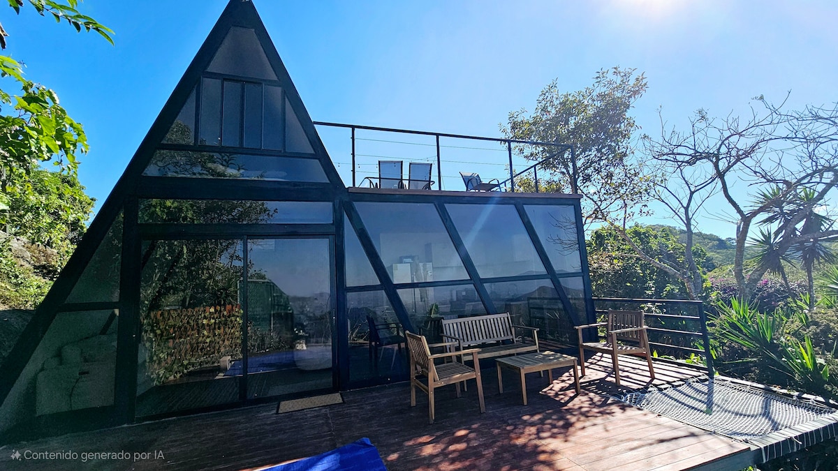 A modern A-frame cabin features large glass panels that allow natural light to fill the interior. The spacious deck showcases several outdoor seating options, including wooden benches and lounge chairs, complemented by the surrounding greenery and panoramic views.