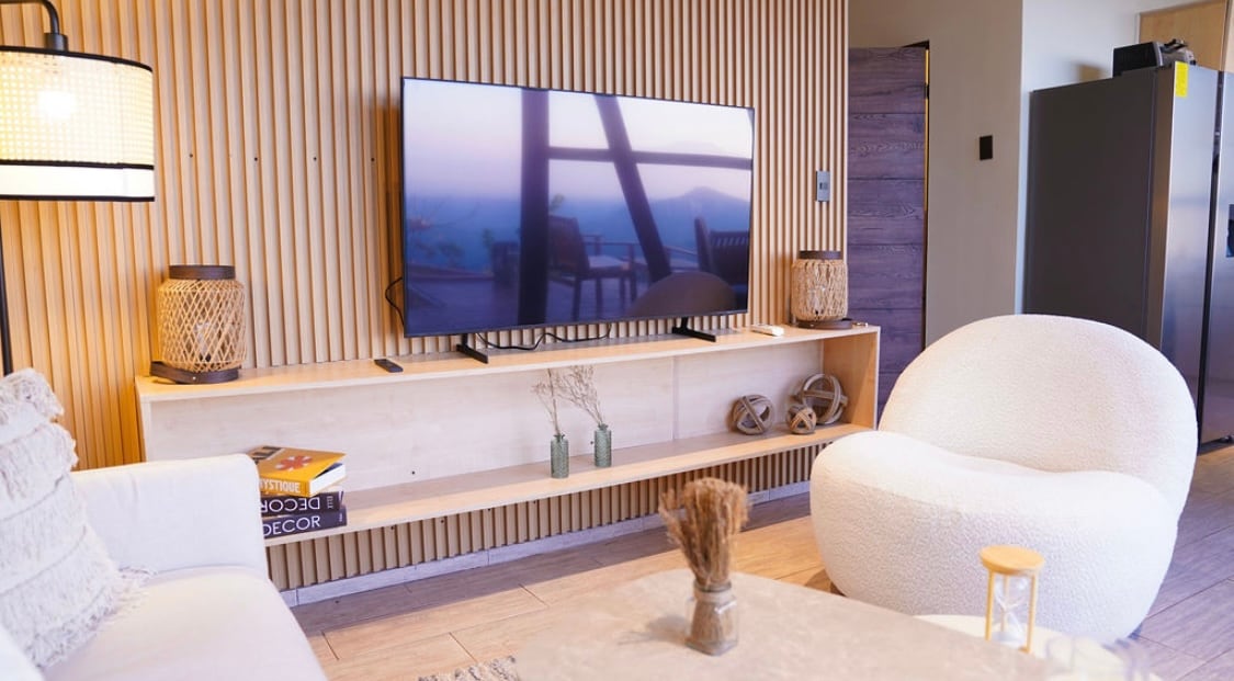 A modern living area showcases a large flat-screen television mounted on a wooden wall, complemented by decorative light fixtures and natural elements. A unique round chair and a low coffee table enhance the inviting space, which benefits from neutral tones and a serene atmosphere.