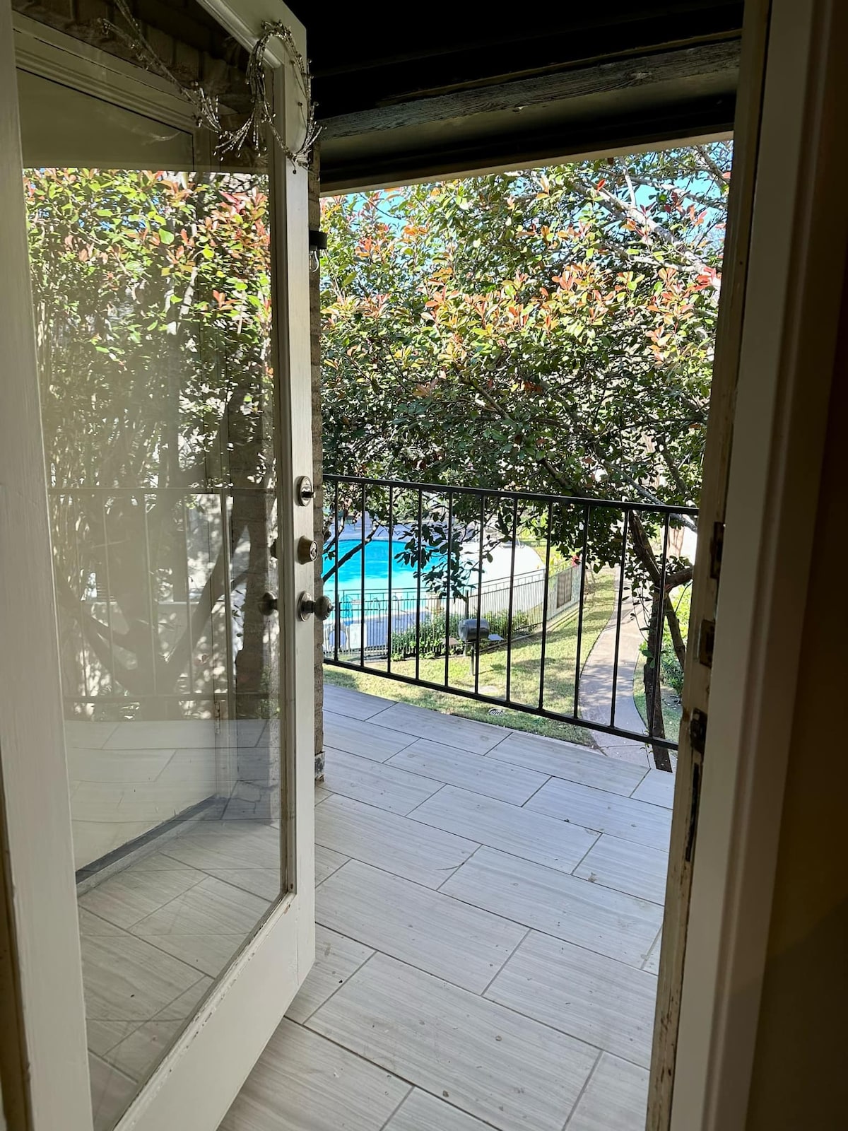 A door is open to a private balcony, framed by a railing overlooking a serene pool surrounded by lush greenery. Sunlight filters through the leaves, creating a bright and inviting atmosphere.