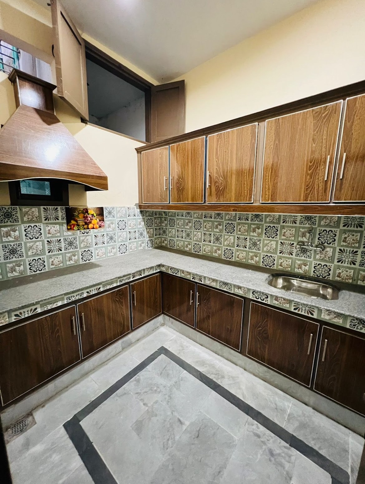 A modern kitchen features wooden cabinetry and a patterned tiled backsplash. A spacious countertop is complemented by a stainless steel sink. Natural light enters through a window, enhancing the clean lines and functional design of the cooking space.